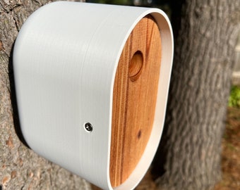 May include: A white birdhouse with a wooden front panel and a round hole for birds to enter. The birdhouse is mounted on a tree trunk.