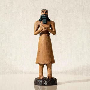 May include: A wooden statue of a figure with a long beard and skirt. The figure has large eyes and is standing on a black base. The statue is a light brown colour.