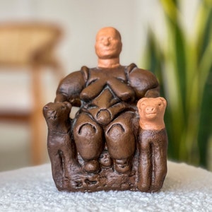 May include: A brown clay figurine depicting a seated figure with a light tan head, flanked by two smaller animal figures on either side. The central figure has a rounded torso and legs, while the two animals have light tan heads and brown bodies.