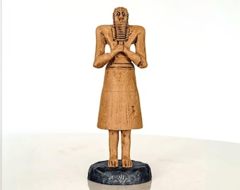 May include: A tan-colored figurine of a standing figure with a long beard and skirt, hands clasped in front. The figure stands on a dark, textured base. The statue has a detailed face and is likely a decorative item.