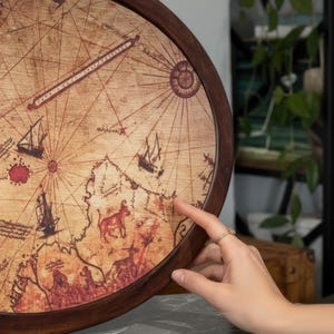 May include: A round wooden wall art piece featuring a vintage map design. The map is printed on a light brown surface with dark brown lines and illustrations of ships and animals. The artwork is framed with a dark brown wooden border.