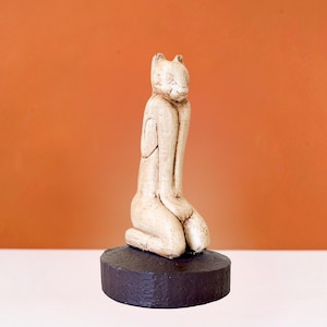 Peut inclure: Figurines de chat en céramique blanche assise sur une base en bois brun foncé. Le chat est en position assise, les pattes repliées sous le corps. La figurine est un design simple et abstrait.