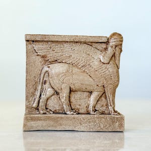 Op de afbeelding: Een beige stenen sculptuur van een gevleugelde leeuw met een menselijk hoofd. Het beeld is gedetailleerd en heeft een getextureerd oppervlak.