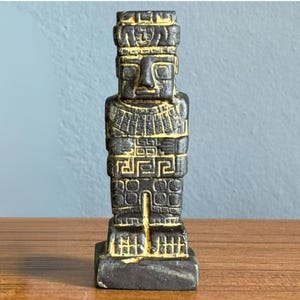 May include: A dark gray statue with gold accents, depicting a stylized human figure. The statue has a rectangular base and is adorned with geometric patterns and a headdress. The figure's face is carved with simple features.
