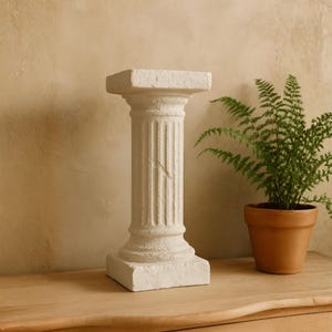 May include: A small, off-white, fluted column with a square top and base. The column has a weathered appearance and sits on a wooden surface. A potted fern is to the right of the column. The background is a light, textured wall.
