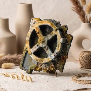 May include: A rectangular decorative object featuring a prominent circular gear-like element with four spokes. The surface has a weathered, textured finish in shades of dark gray, muted green, and rusted orange, resembling aged metal or stone.