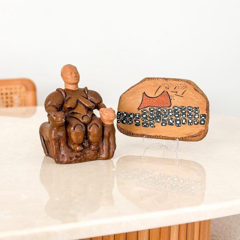 May include: A brown clay sculpture of a seated figure flanked by two small animals, positioned next to a flat, irregularly shaped brown clay plaque featuring a painted orange triangular shape and a grid of black and white rectangular patterns.