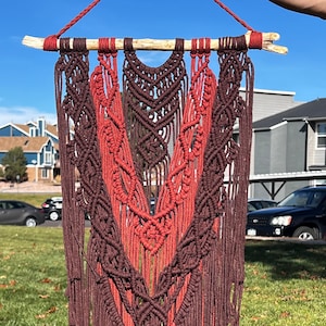 Puede incluir: Un tapiz de macramé de pared con un diseño en forma de V en tonos marrones y rojos. El tapiz está hecho con cordón de algodón y tiene una barra de madera natural en la parte superior.