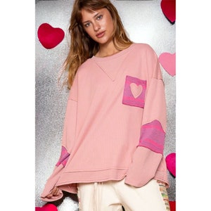 Puede incluir: Una sudadera rosa de manga larga con un parche en forma de corazón en el bolsillo y tejido a rayas rosa y morado en las mangas.