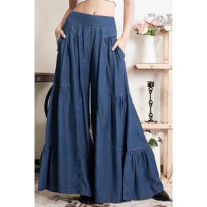 May include: Wide-leg denim pants with a tiered ruffle design. The pants are a dark blue wash and have a high waist.