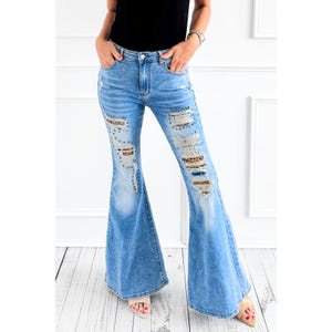 May include: Light blue denim bell-bottom jeans with distressed details. The jeans feature ripped sections with leopard print patches and are embellished with small silver studs. The jeans have a classic five-pocket design.