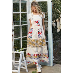 May include: Cream-colored overall dress with floral patchwork details. The dress features red and pink roses, and a tiered skirt with a patterned design. The model is wearing a white t-shirt and sneakers.