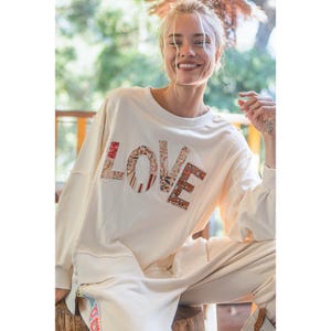 May include: A white long-sleeved sweatshirt with a large patchwork "LOVE" graphic on the front. The sweatshirt is made of a soft, comfortable fabric and has a relaxed fit.