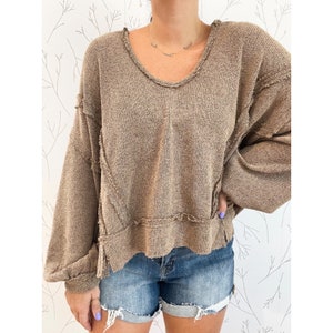 May include: A brown, long-sleeved sweater with a distressed, frayed hem and a relaxed fit. The sweater has a scoop neck and a slightly cropped length.
