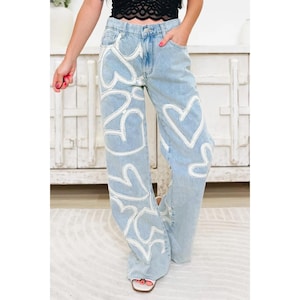 Op de afbeelding: Lichtblauwe jeans met wijde pijpen en een wit hartpatroon op de voorkant. Het model draagt witte sandalen. De jeans heeft zakken en een knoop- en ritssluiting.