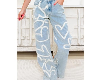 May include: Light blue denim jeans with a wide leg design. The jeans feature a white heart pattern throughout the front of the legs. The model is wearing white sandals. The jeans have pockets and a button and zipper closure.