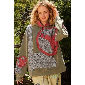 May include: Olive green hoodie with a patchwork design. The hoodie features a red peace sign on the front and sleeve, and floral patterned fabric. The hood and accents are red. The model is wearing the hoodie.