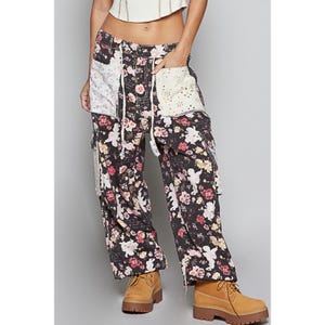 May include: Black floral print cargo pants with white lace pocket details and a drawstring waist. The pants feature a mix of pink, white, and yellow flowers on a dark background. The pants are paired with tan work boots.