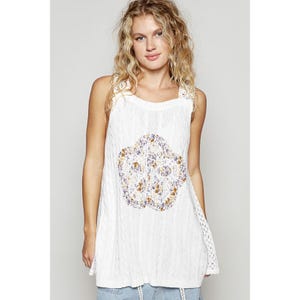 May include: White tank top with a floral design. The top features a textured knit fabric with a round neckline and lace detailing on the straps and sides. The floral design is in the center of the top and has a mix of purple, yellow, and white colors.
