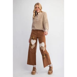Puede incluir: Una mujer lleva un suéter beige con cuello alto y cierre de botones, combinado con pantalones anchos marrones con corazones de patchwork en varios tonos de marrón, blanco y cuadros.