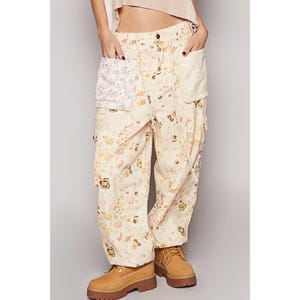 May include: Cream-colored cargo pants with a floral pattern in pink, yellow, and green. The pants have multiple pockets and are paired with tan work boots. The pants have a relaxed fit and a high waist.