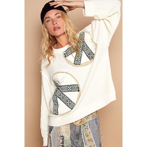Puede incluir: Sudadera blanca de manga larga con dos símbolos de paz bordados. Los símbolos de paz están hechos de cinta negra y blanca y están rodeados de un círculo beige.