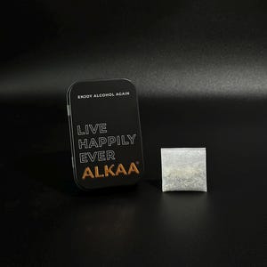 May include: A black rectangular tin with the text "ENJOY ALCOHOL AGAIN" and "LIVE HAPPILY EVER ALKAA" in gold lettering. A small, square, silver packet sits to the right of the tin. The background is black.