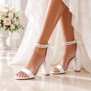White bridal block heel sandals with double pearl ankle straps and ribbon bows, worn by a bride under a lace wedding dress.