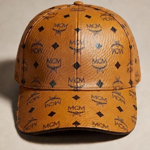 May include: A brown baseball cap featuring a repeating pattern of the letters "MCM" and a logo. The cap has a curved peak and a rounded crown. The cap is made of a leather-like material.