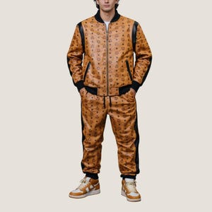 Puede incluir: Conjunto de chándal marrón a juego, compuesto por chaqueta y pantalón. La chaqueta tiene cremallera, ribetes negros y detalles negros en los hombros. Los pantalones tienen rayas negras en los laterales. El conjunto tiene un patrón de logotipo repetido.