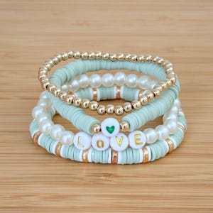 May include: Stack of four bracelets. The bracelets feature a mix of gold beads, white faux pearls, and mint green disc beads. One bracelet spells out the word "LOVE" with white letter beads and a green heart.