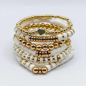 May include: Stack of six white and gold beaded bracelets. The bracelets feature gold beads, white disc beads, and a green heart charm. The bracelets are stacked on top of each other against a white background.