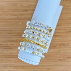 May include: A stack of seven beaded bracelets featuring white and gold-colored beads. The bracelets include letter beads spelling JESUS and JOY, heart-shaped beads, and a small gold-colored cross charm. The bracelets are displayed on a white cylindrical stand.
