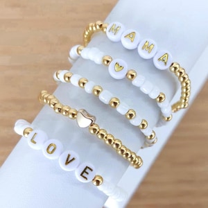 May include: Five white beaded bracelets with gold beads and gold lettering. The bracelets spell out "LOVE" and "MAMA" with a heart charm on each.