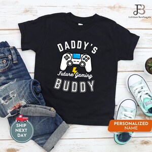 Può includere: Maglietta nera con un'immagine di un controller per videogiochi bianco e il testo "Daddy's Future Gaming Buddy".