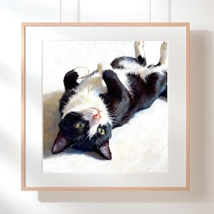 May include: A framed square art print featuring an oil painting of a black and white tuxedo cat lying on its back. The cat has green eyes, white paws, and a white chest, positioned against a bright, light-colored background with soft shadows.