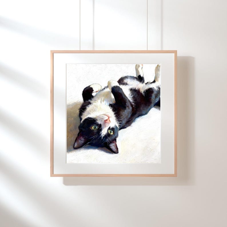 May include: A framed square art print featuring an oil painting of a black and white tuxedo cat lying on its back. The cat has green eyes, white paws, and a white chest, positioned against a bright, light-colored background with soft shadows.