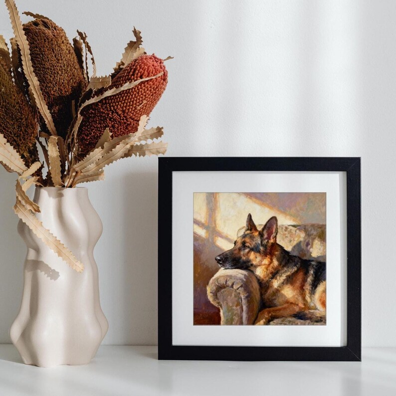 The German Shepherd print in a black frame displayed on a white shelf beside a cream-colored ceramic vase with brown dried stems.