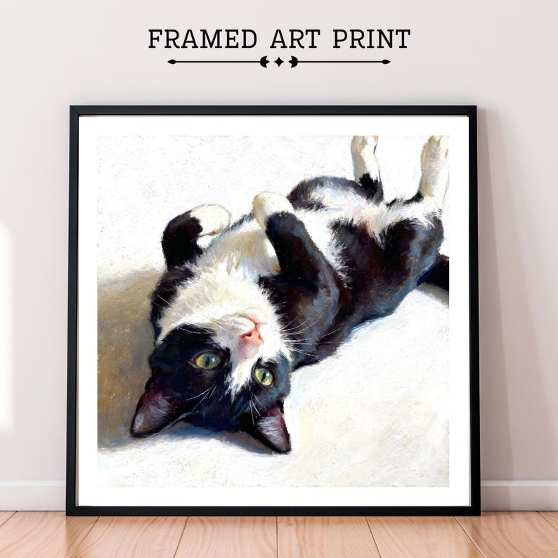 A framed oil pastel art print of a black and white tuxedo cat lying on its back with green eyes, looking up at the viewer.