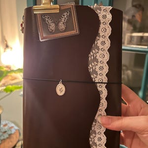 May include: A dark brown leather journal featuring a scalloped edge, adorned with a white lace trim along the side. A gold metal clip holds a small rectangular card with a butterfly illustration, and a black elastic band secures a silver oval charm.