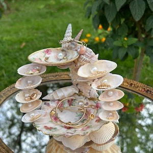 Könnte beinhalten: Ein mehrstufiger Schmuckständer aus verschiedenen Muscheln mit zwei kleinen Porzellanschalen mit Blumenmuster. Die Struktur hält mehrere goldfarbene Ringe, Ohrringe und Halsketten, wobei kleine weiße Perlen die Muscheln akzentuieren.