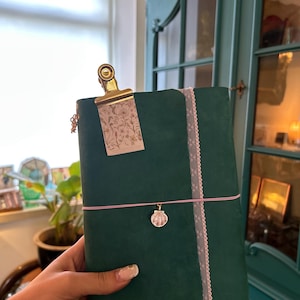 Könnte beinhalten: Ein dunkelgrünes Notizbuch mit matter Oberfläche, das auf der rechten Seite mit einem vertikalen Streifen aus weißer Spitze verziert ist. Ein goldener Metallclip hält oben ein florales Etikett, und ein dünnes rosa Gummiband sichert einen Muschelanhänger.