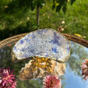 Puede incluir: Un plato decorativo de concha de ostra con interior blanco, motivos florales azules y borde pintado en oro. La concha descansa sobre un pequeño soporte dorado y contiene un collar de cuentas transparentes facetadas con una perla blanca.