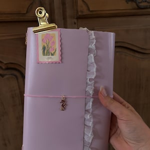 Könnte beinhalten: Ein hellvioletter Notizbuchumschlag mit weißer, gerüschter Spitzenborte an der rechten Kante, einer goldenen Metallklemme oben und einem rosa Gummiband mit einem kleinen goldenen Seepferdchen-Anhänger. Ein rechteckiger Aufkleber mit rosa Tulpen und dem Text TULIPS ist angebracht.