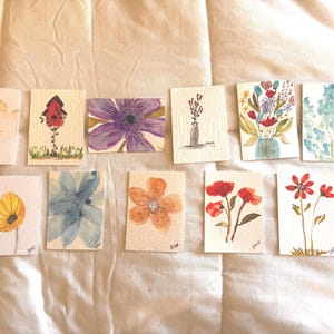 Könnte beinhalten: Eine Sammlung von Aquarell-Blumenkunstkarten. Jede Karte zeigt ein einzigartiges Blumendesign in verschiedenen Farben, darunter Lila, Orange, Rot und Blau. Einige Karten zeigen einzelne Blumen, während andere Blumensträuße oder Blumenarrangements zeigen. Die Karten sind auf einer weißen Oberfläche angeordnet.