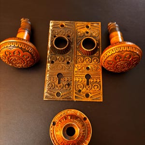 May include: Antique door hardware set featuring two ornate door knobs, two rectangular backplates with keyholes, and a round escutcheon. The hardware is made of a warm, aged brass with detailed floral and geometric patterns.