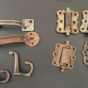 Op de afbeelding: Een assortiment vintage metalen hardware, waaronder scharnieren, grendels en haken, in verschillende vormen en maten. De stukken hebben een verweerde, verouderde uitstraling met tinten bruin, grijs en groen, wat suggereert dat ze antiek of gerecycled zijn.