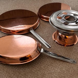 Può includere: Un set per fonduta in rame con un supporto per bruciatore in acciaio inossidabile e due manici. Il set include tre piatti in rame e una pentola per fonduta in rame. Gli articoli sono disposti su un tappeto marrone.