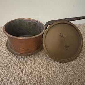 Peut inclure: Une marmite en cuivre antique avec son couvercle. La marmite est de forme ronde avec une base large et un intérieur sombre. Le couvercle est d'une couleur plus claire et possède une poignée. La marmite est posée sur une surface texturée.