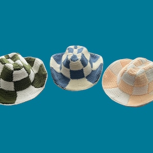 Op de afbeelding: Drie gehaakte hoeden met een geruit patroon in groen, blauw en perzikkleur. De hoeden hebben een brede rand en zijn gemaakt van wit garen.